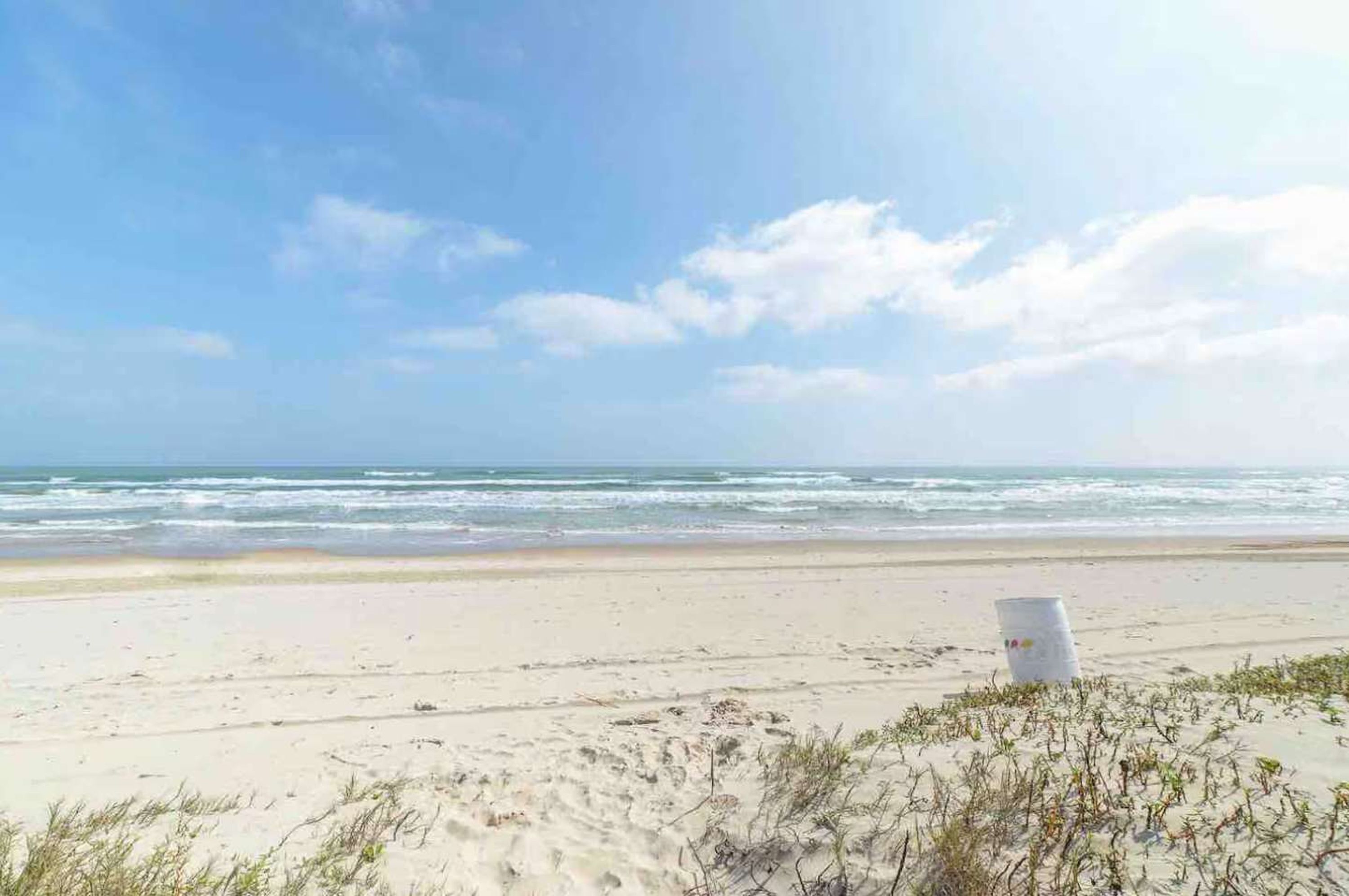 A beach with a white bucket and sparse vegetation in the foreground.