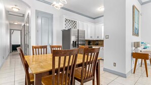 A modern kitchen with white cabinets, a stainless steel refrigerator, and a dining area with wooden chairs and a table. - Modern Beachside Condo w Pool - South Padre Island (South Padre Island)