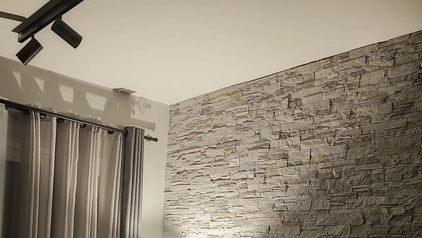 Ein modernes Wohnzimmer mit einer Wand im Steinoptik, einem grauen Sofa, einem Holztisch und einer Stehlampe.