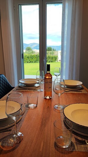 A dining table with plates, glasses, and a bottle of wine, set against a backdrop of a scenic outdoor view through large windows. - Alaia Kefalonia Garden Haven Elegant Holiday Home (Kefalonia)
