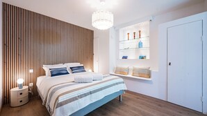 Ett modernt sovrum med en väggpanel av träribbor, en säng med randigt sänglinne, ett sängbord med en lampa och en inbyggd hylla med dekorativa föremål.
