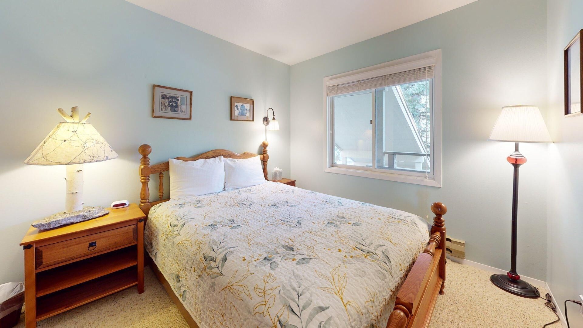 A bedroom with a bed, bedside lamps, a nightstand, a window, and wall-mounted pictures.