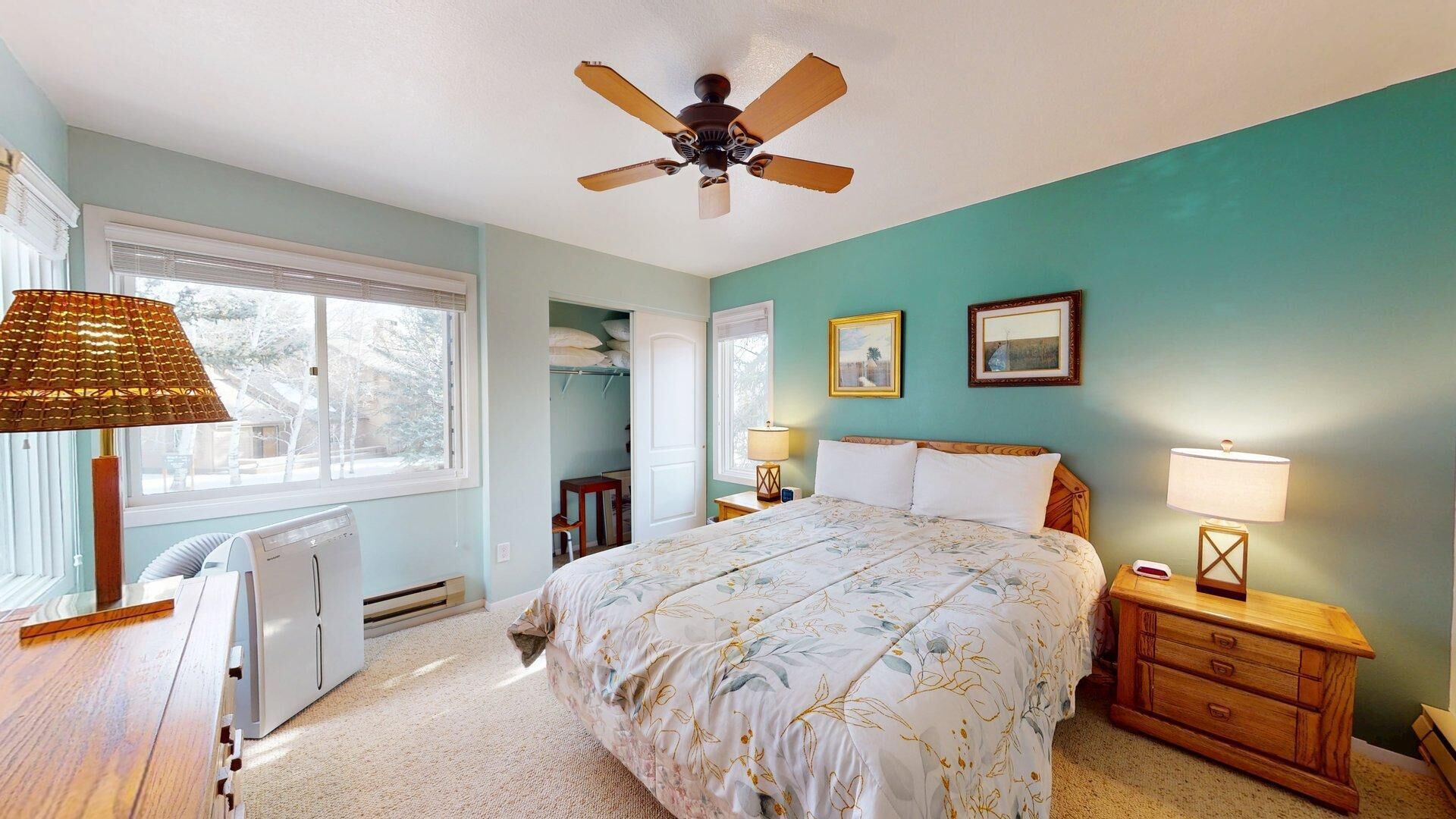 A bedroom with a bed, a ceiling fan, a window with blinds, a radiator, a desk, and a lamp.