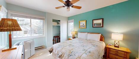 A bedroom with a bed, a ceiling fan, a window with blinds, a radiator, a desk, and a lamp.