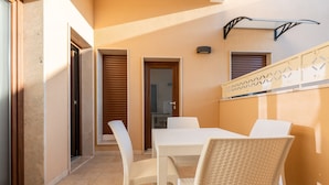 Une terrasse ensoleillée avec une table blanche et des chaises en osier, une porte et une étagère fixée au mur.