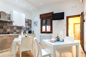 Una cucina moderna con mobili bianchi, un tavolo da pranzo con sedie, un televisore a schermo piatto e un rivestimento a parete in piastrelle decorate.
