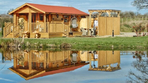 A wooden cabin with a red roof, a porch, and a small adjacent structure with a circular window. - The Balmoral Studio Lodge – Private Hot Tub & On-Site Carp Fishing 🎣🛁 (Ellesmere)