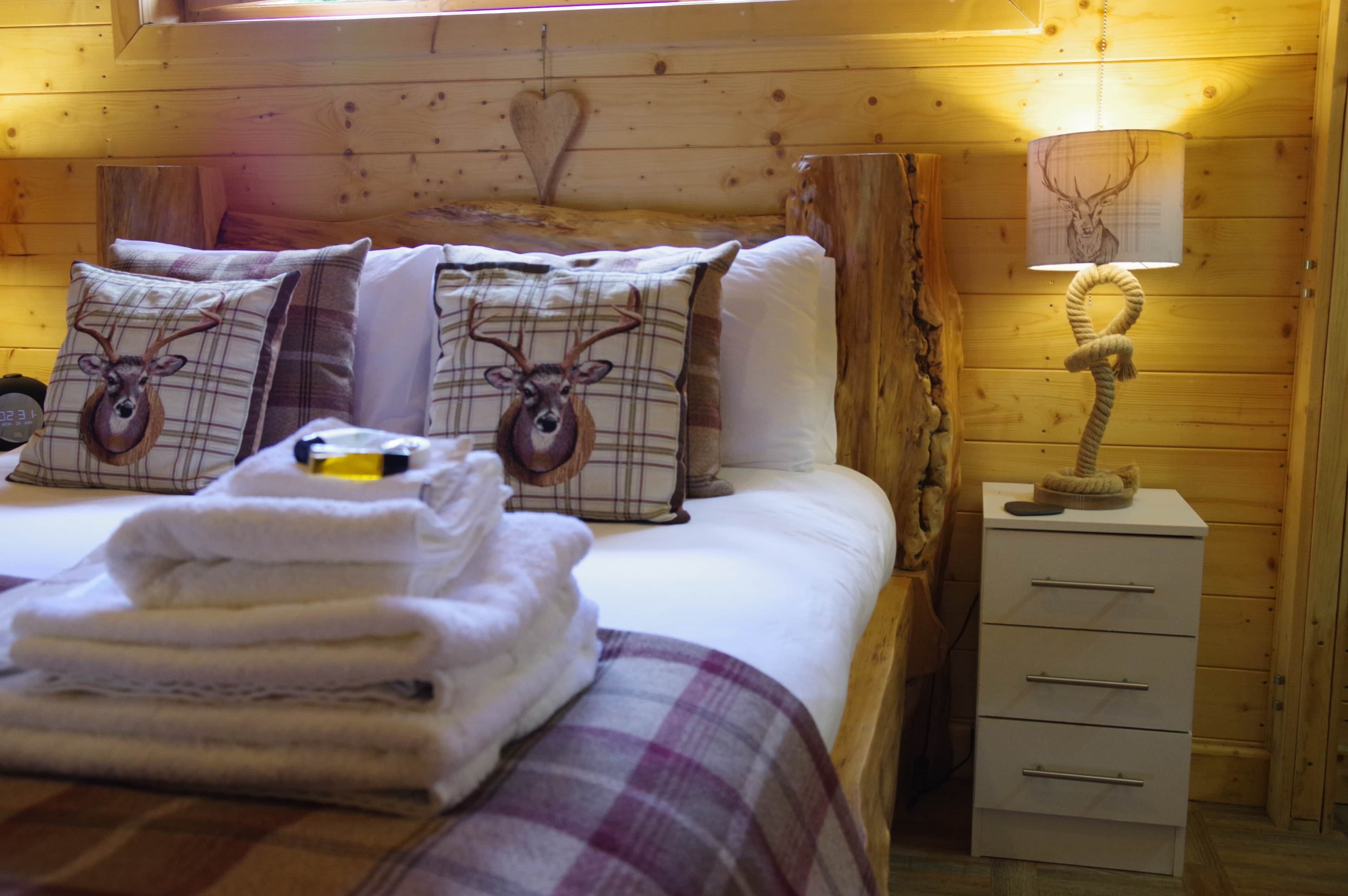 A wooden cabin room with a bed, pillows with deer designs, a lamp, and a stack of towels.