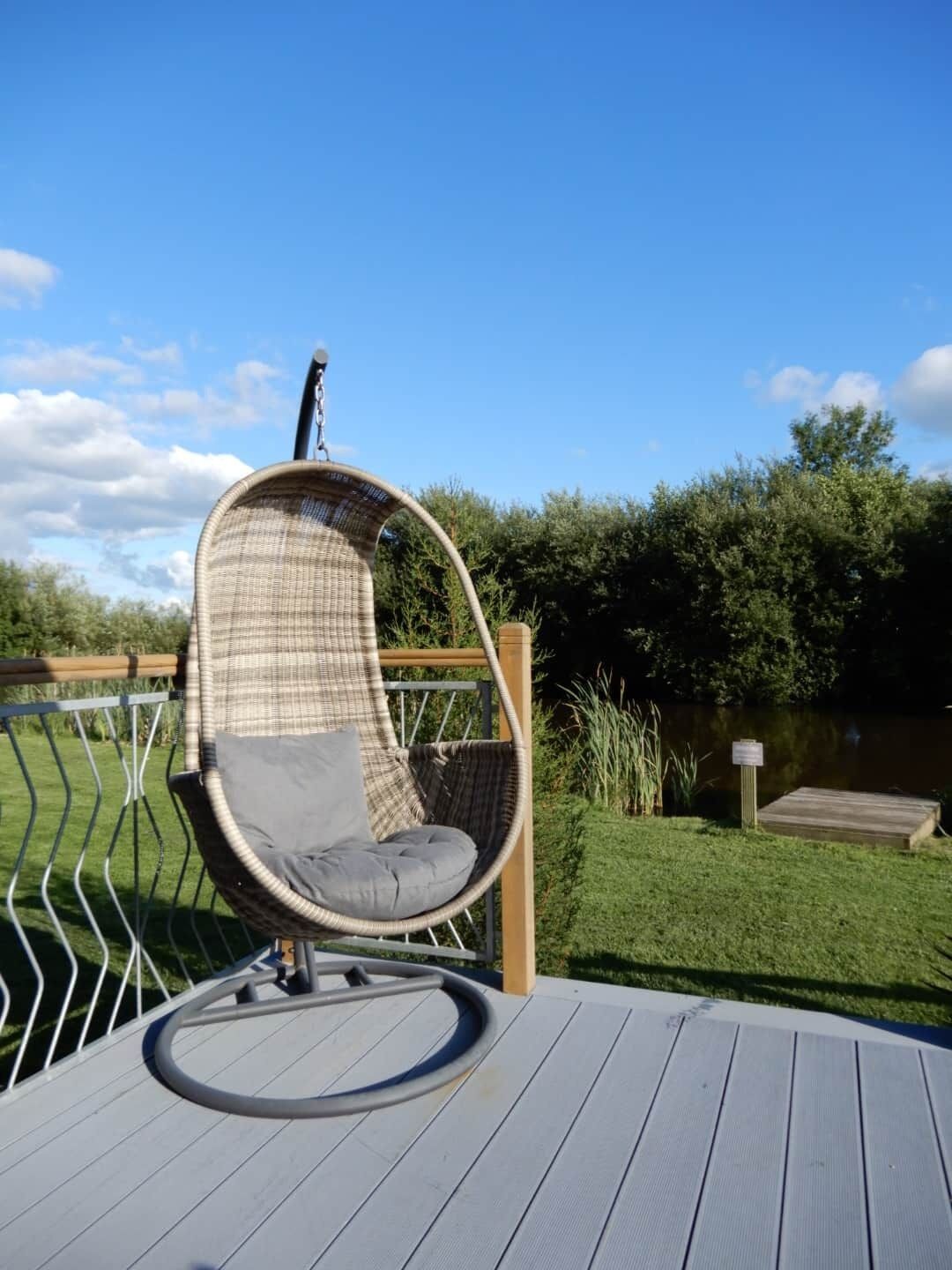 Ein hängender Korbstuhl mit Kissen auf einer Holzterrasse mit Blick auf eine Rasenfläche und ein Gewässer.
