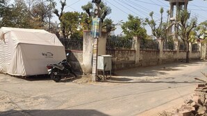 A covered motorcycle, a utility box, and a covered vehicle on a street with a fence and trees in the background.