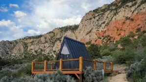 A cabin with a metal roof and wooden deck, surrounded by desert vegetation and a dirt path leading to it.