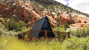 A mountain cabin with a wooden deck and a metal railing, surrounded by natural vegetation and a rocky hillside.