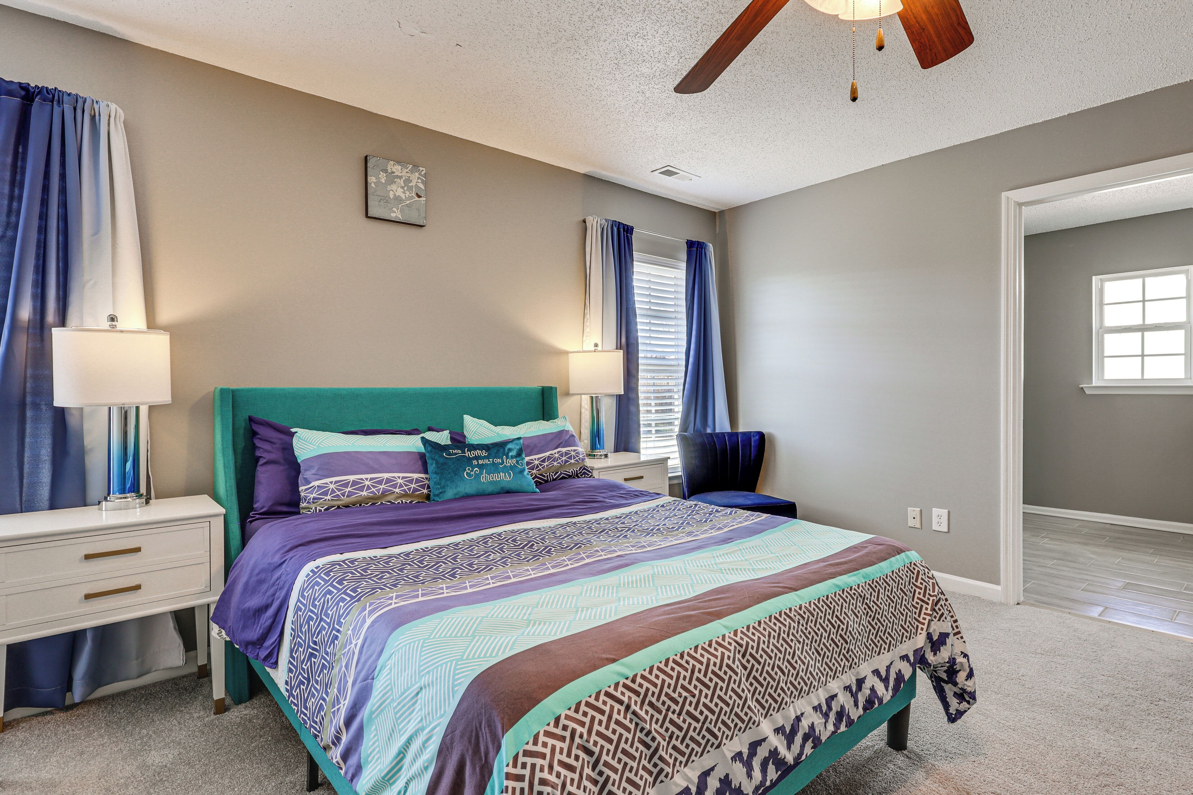 A bedroom with a bed, two bedside tables, a chair, a ceiling fan, and a window with curtains.