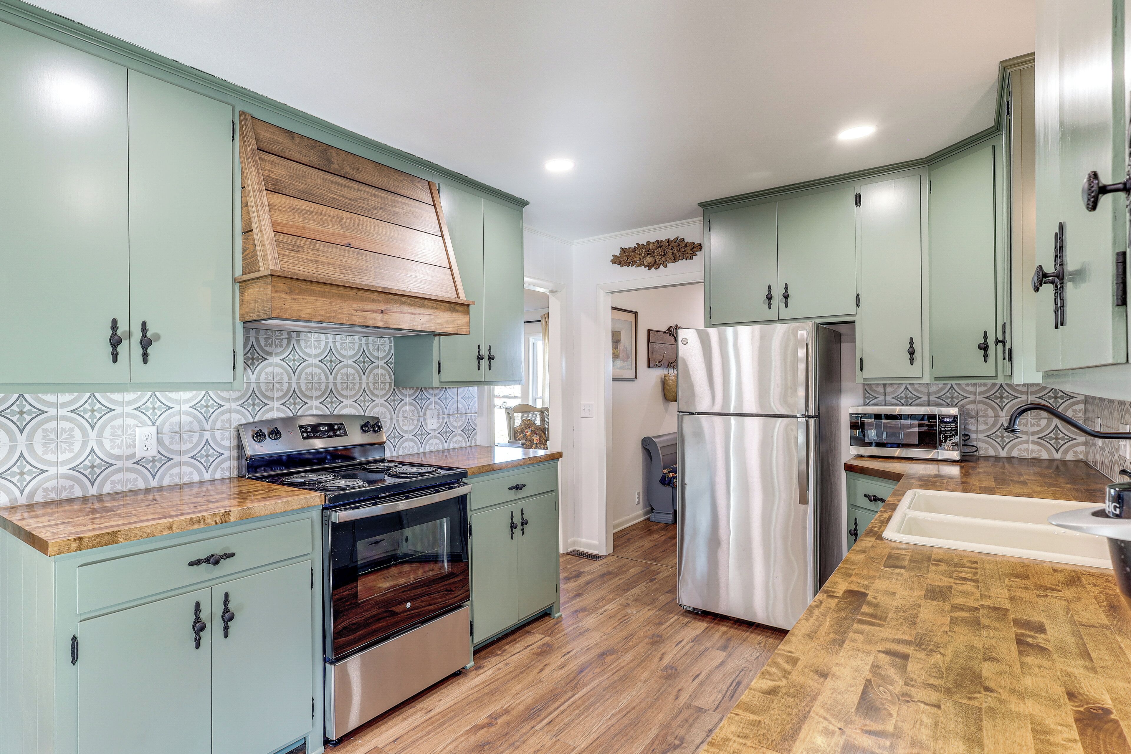 A kitchen with light green cabinets, a wooden hood, stainless steel appliances, and a patterned backsplash.
