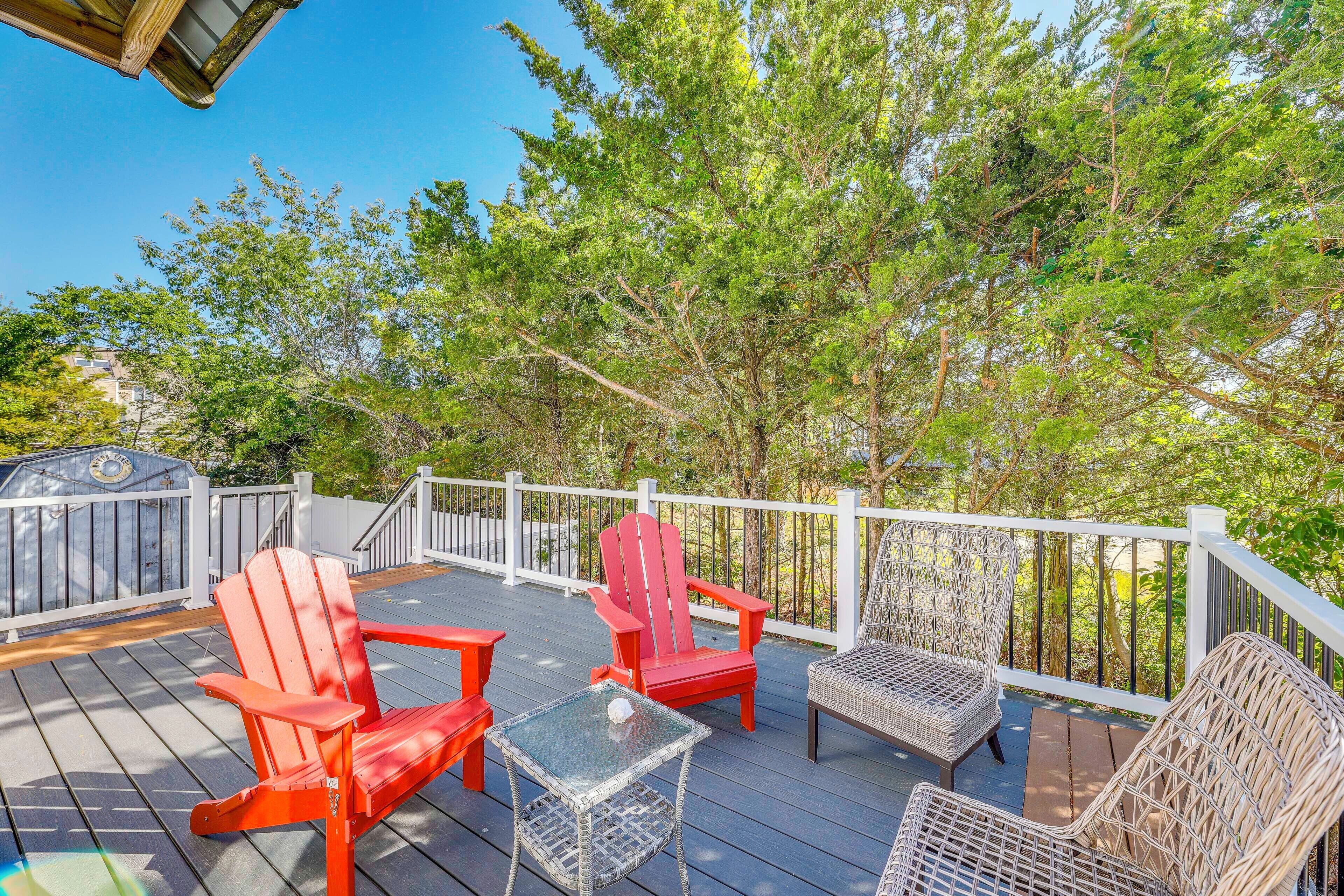 A deck with outdoor seating, including red Adirondack chairs and wicker chairs, surrounded by trees.