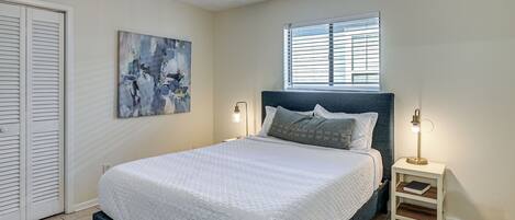 Een slaapkamer met een bed, een nachtkastje, een plafondventilator, een raam met jaloezieën en een schilderij aan de muur.