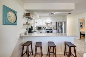 A modern kitchen with white cabinets, a stainless steel refrigerator, a microwave, and a large island with bar stools. - Steps to Sea! Panama City Gem w/ Pet-friendly Yard (Panama City Beach)