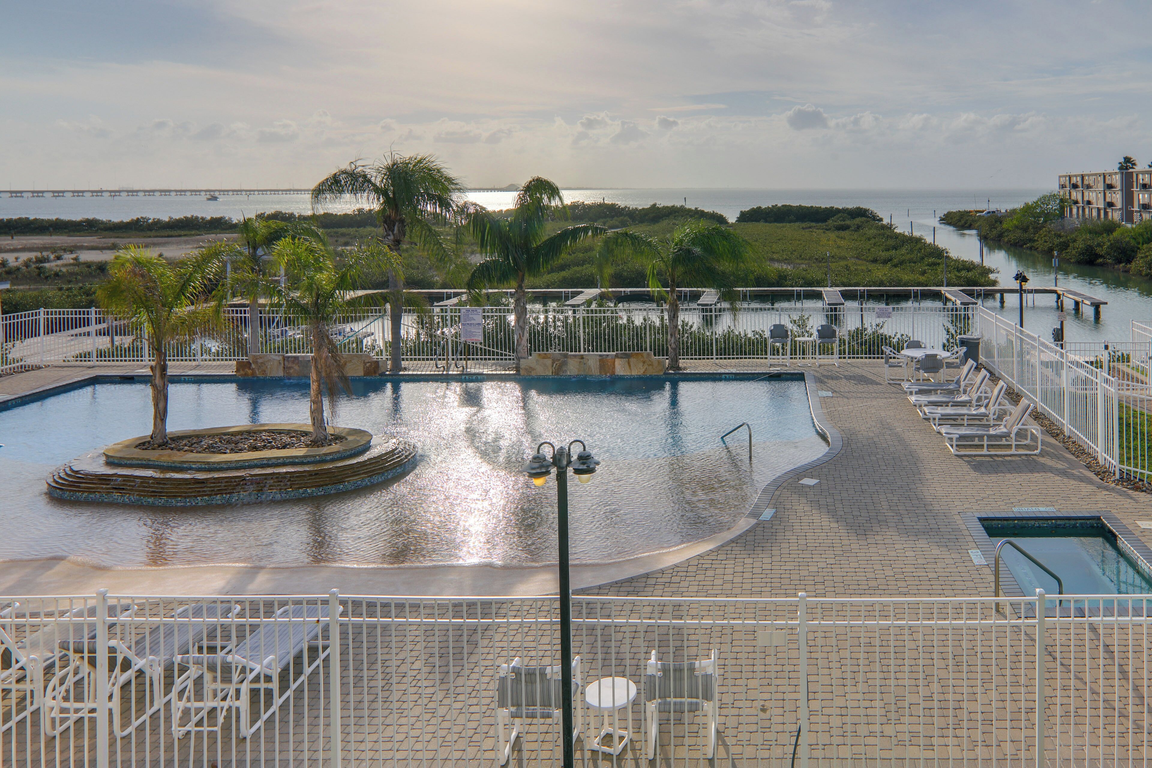 Zona de piscina con vista al agua, palmeras y un puente al fondo.