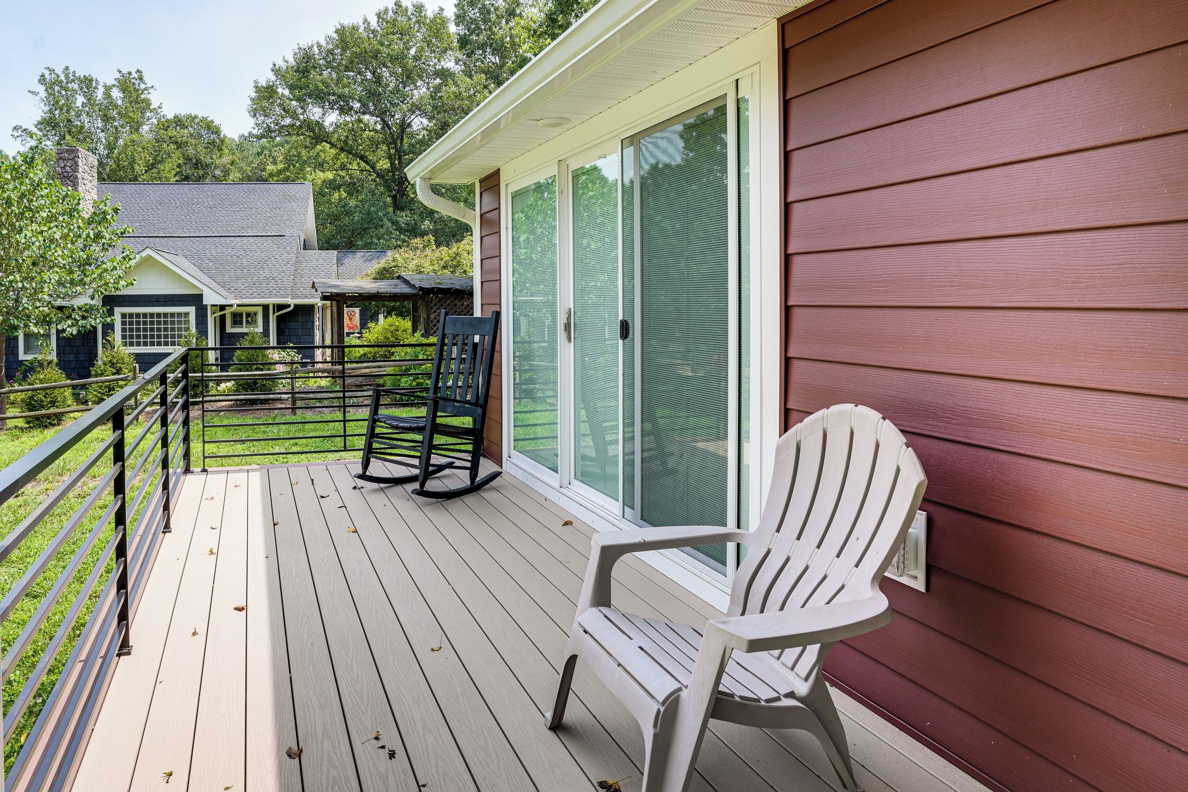 A wooden deck with a white chair and a rocking chair, a glass door, and a view of a house with a chimney.