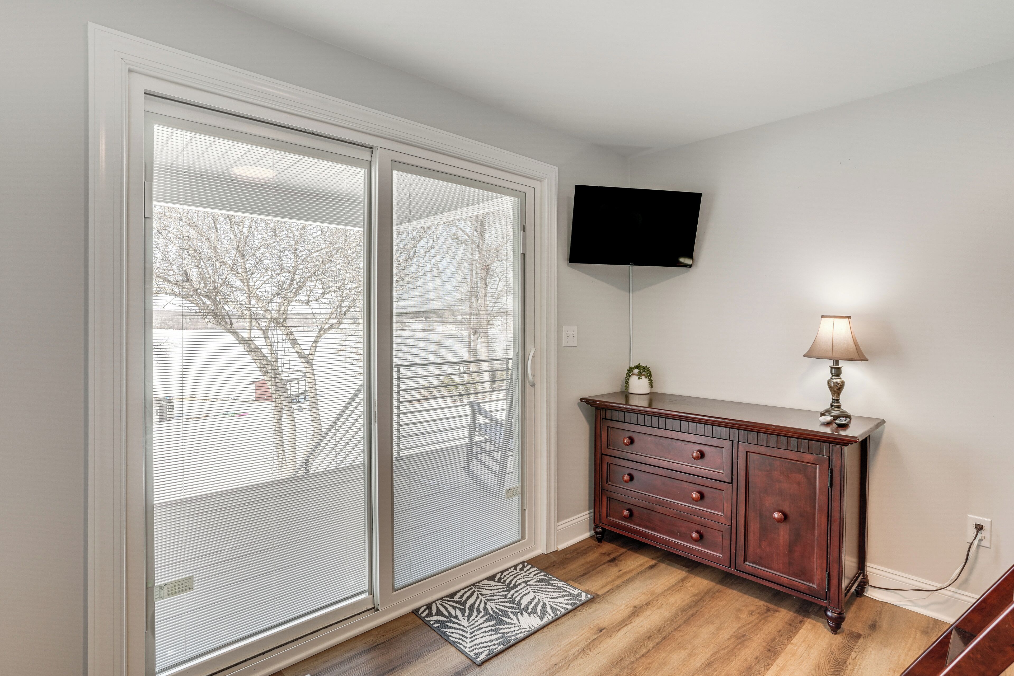 A room with a sliding glass door leading to a balcony, a wooden dresser with drawers, a mounted flat-screen TV, and a floor lamp.