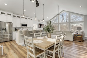 A spacious kitchen with white cabinetry, a central island, and a dining area with a wooden table and white chairs. - Hot Tub + Fire Pits: Waterfront Lake Anna Escape! (Louisa)
