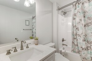 Un baño moderno con un lavamanos blanco, dos llaves, un espejo, una ducha con cortina estampada con motivos florales y un inodoro.