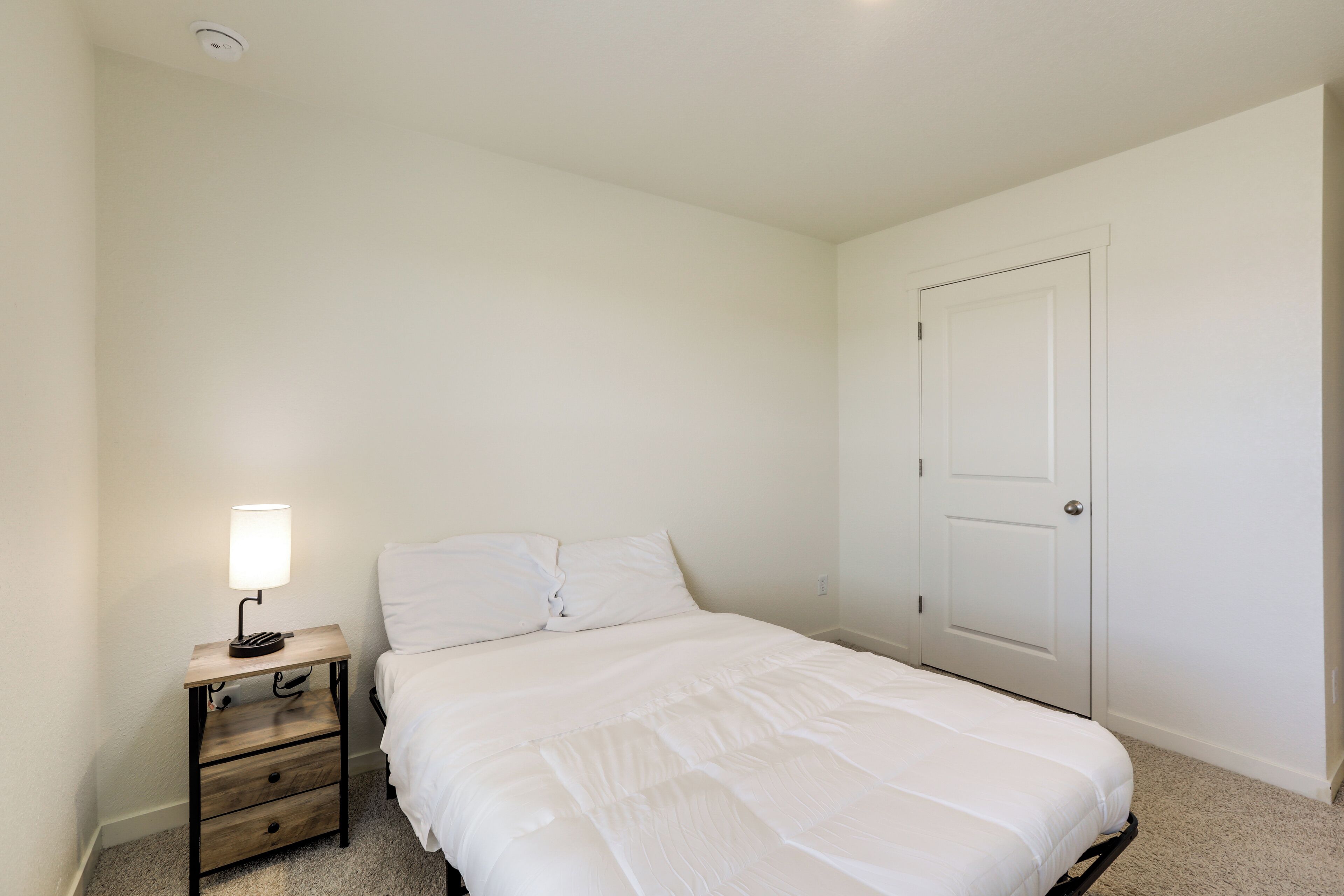 A bedroom with a bed, a nightstand with a lamp, a door, and a carpeted floor.