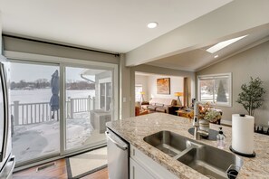 A modern kitchen with a large island, a double sink, and a view of a snowy outdoor area. - Hike, Fish & Golf: Waterfront Gem on Camp Lake! (Sparta)