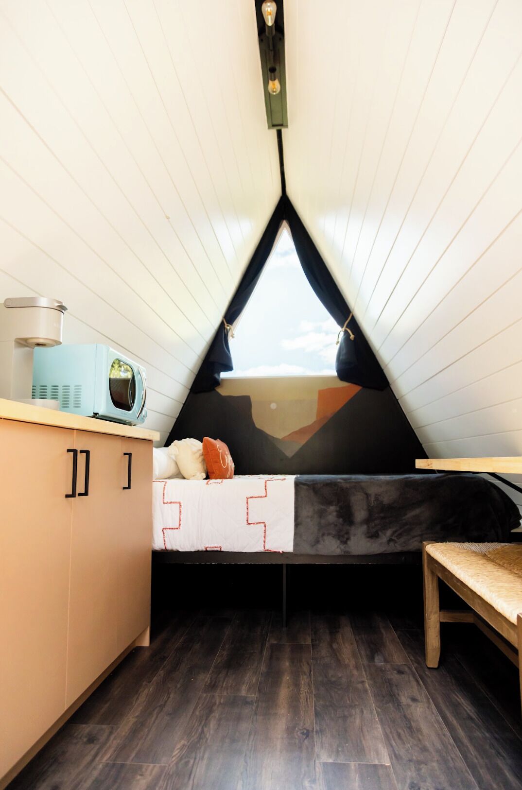 Una habitación pequeña y triangular con cama, mesa de madera, microondas y un armario.