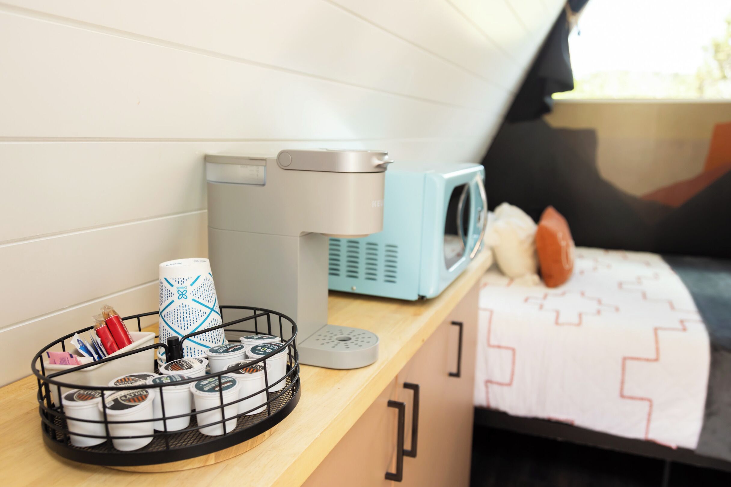 A kitchenette with a coffee maker, microwave, and a basket of coffee pods on a wooden countertop.