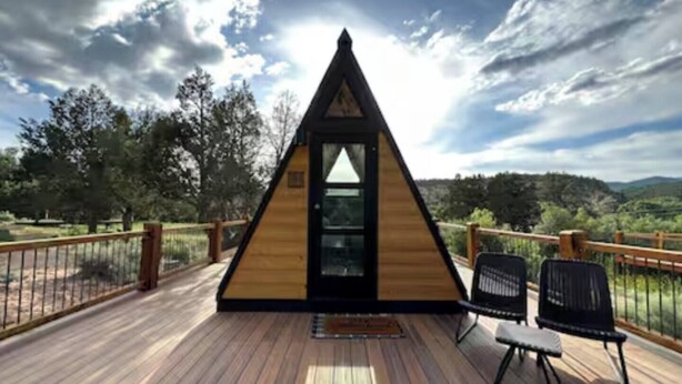 A wooden triangular structure with a glass door, two black chairs, and a wooden deck with a railing. - NEW! Unique Modern Glamping Chalet #3 (Orderville)