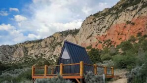 A rustic cabin with a metal roof and wooden deck, surrounded by dry shrubs and a dirt path leading to it. - NEW! Unique Modern Glamping Chalet #3 (Orderville)