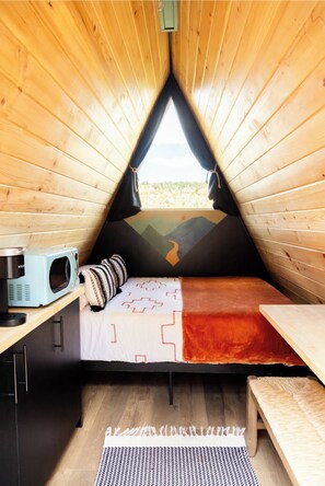 Una tienda de madera triangular y acogedora con una cama, microondas y vistas al exterior.