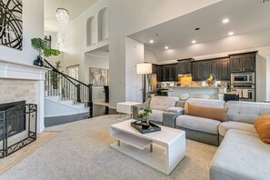 A modern living room with a fireplace, a staircase, and a kitchen area with dark cabinets and stainless steel appliances. - Modern Home w/ Office: Near Parks, Lake & Golf! (Little Elm)