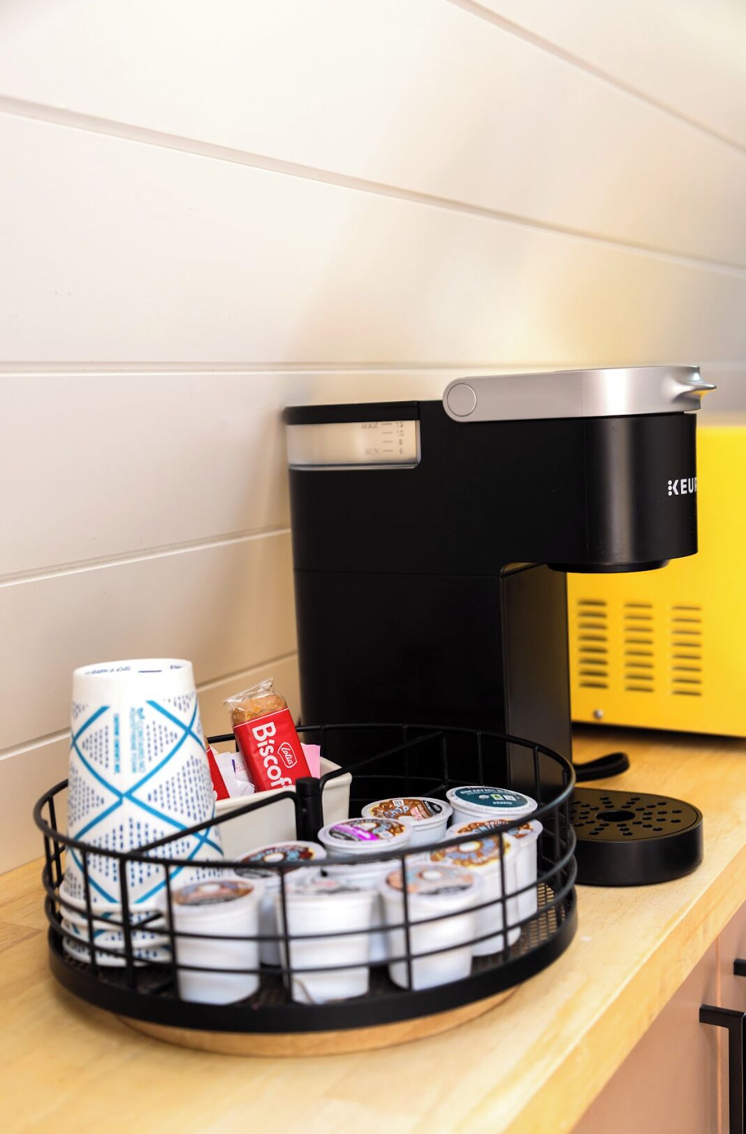 Uma bancada de cozinha com uma cafeteira, um cesto de cápsulas de café e um saco da marca Biscuits.