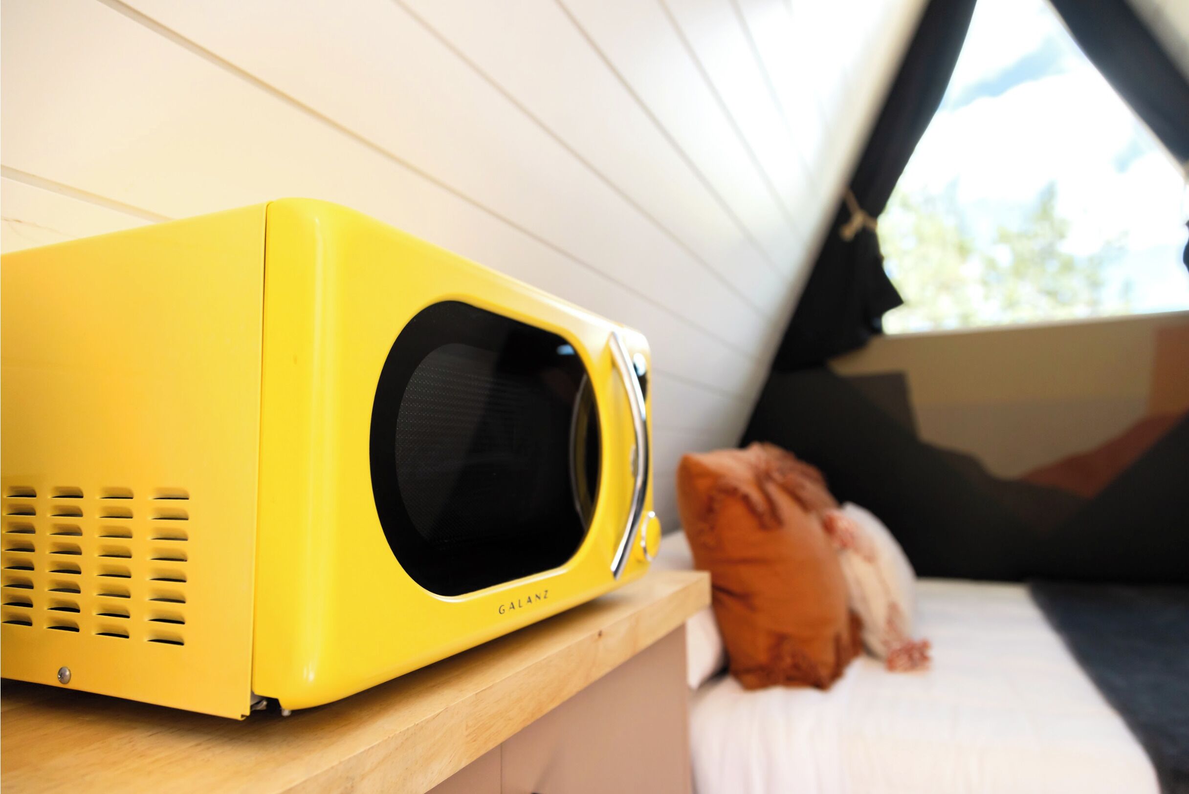 A yellow microwave on a wooden shelf in a room with a bed and a window.
