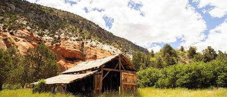 Una estructura de madera rústica con techo inclinado, rodeada de hierba alta y un paisaje montañoso.
