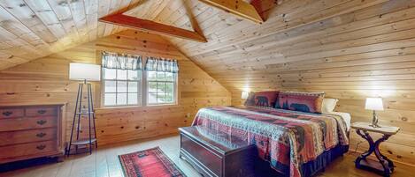 A cozy attic bedroom with a wooden ceiling, a large bed with a colorful blanket, a dresser, a nightstand with a lamp, and a window with curtains.