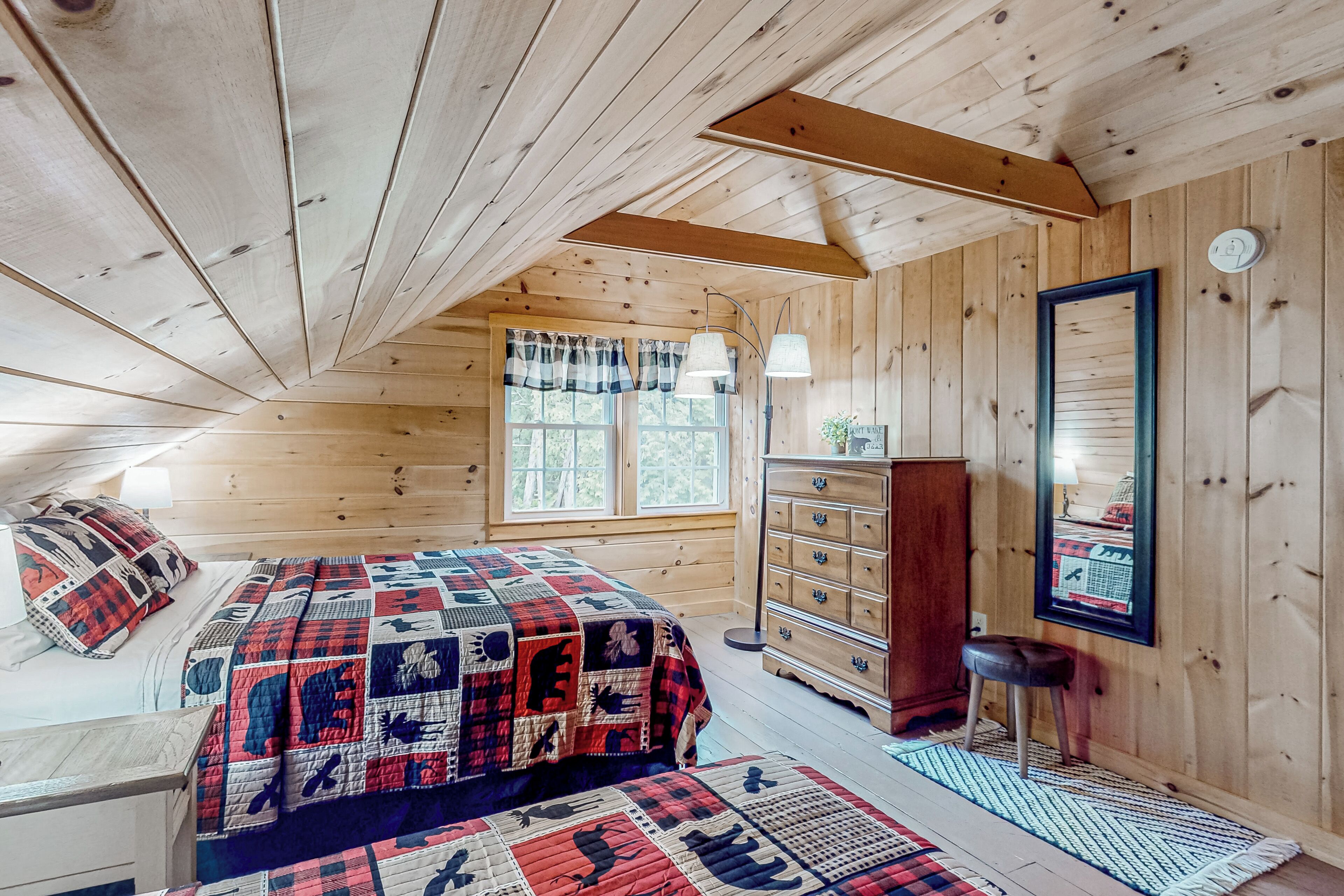 A cozy bedroom with a wooden ceiling, a large bed with a quilt, a dresser, and a mirror on the wall.