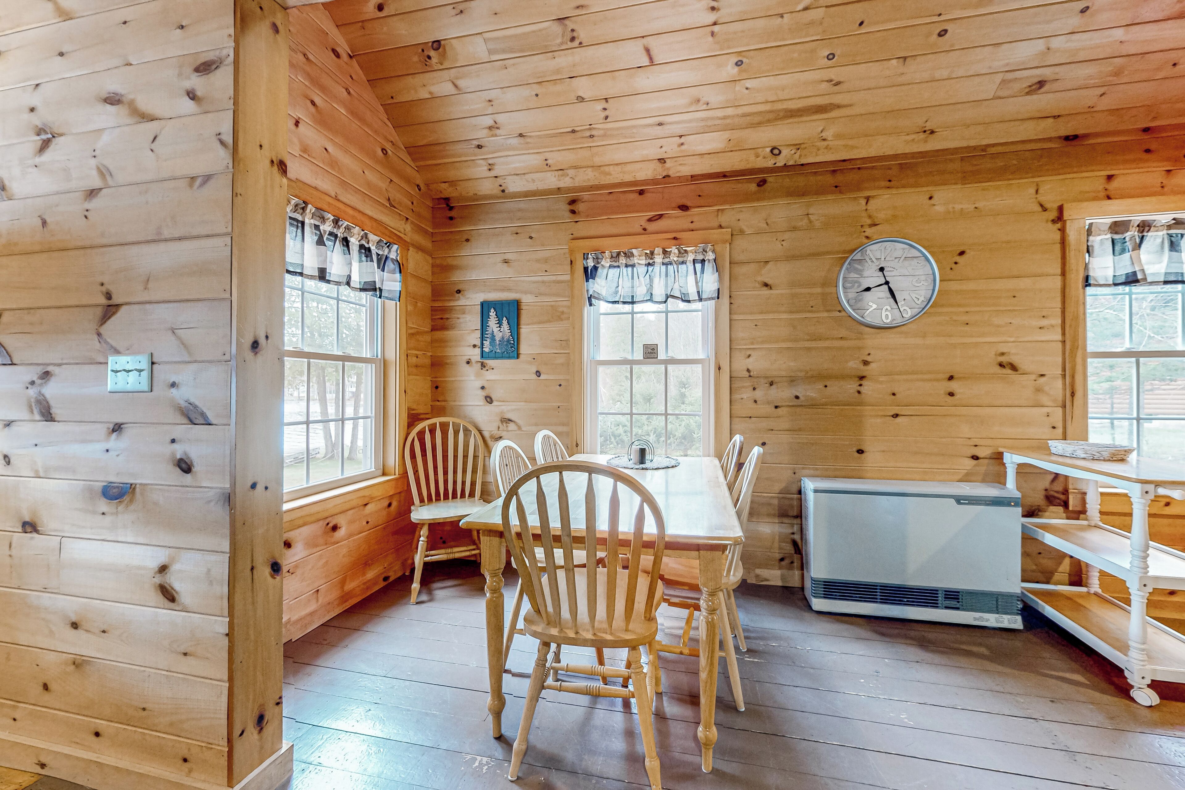 Interior de una cabaña de madera con una mesa de comedor, sillas, un reloj y una ventana con cortinas.