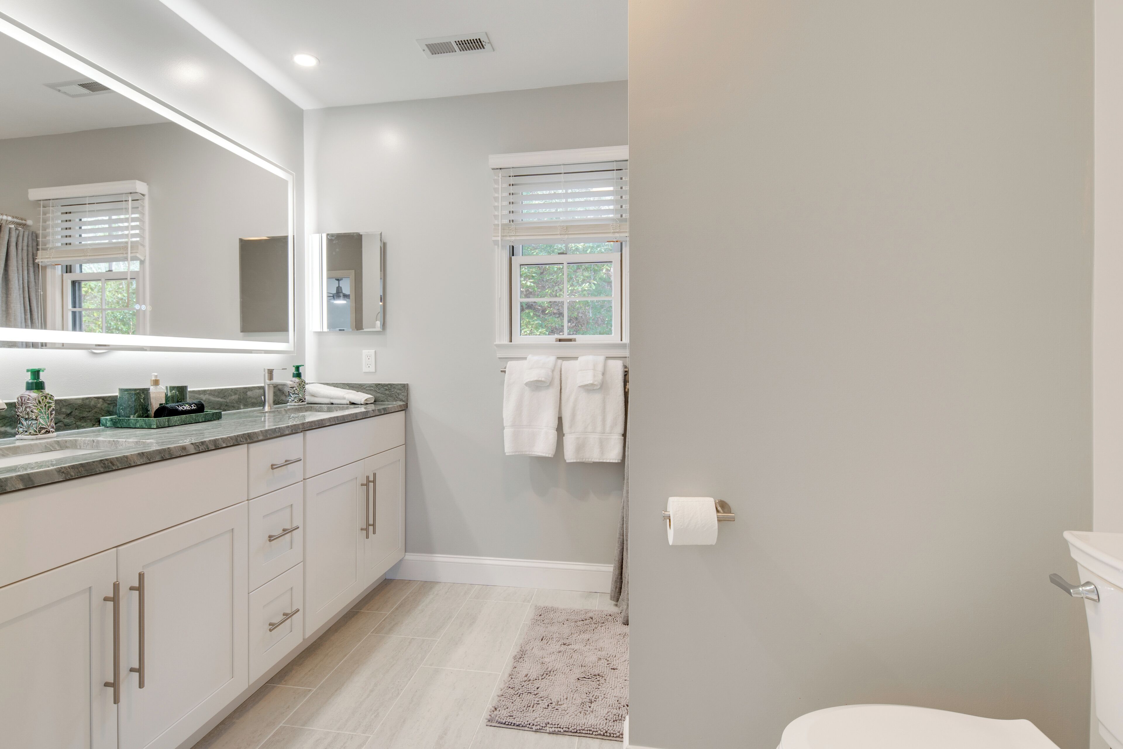 A modern bathroom with a large mirror, white cabinets, a marble countertop, and a toilet.