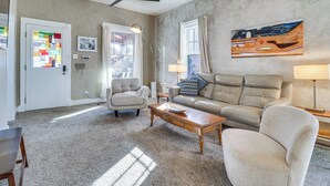A living room with a beige sofa, a wooden coffee table, and a ceiling fan. There is a window with curtains and a painting on the wall. - Deck & Home Office: Walkable Albuquerque Home (Albuquerque)