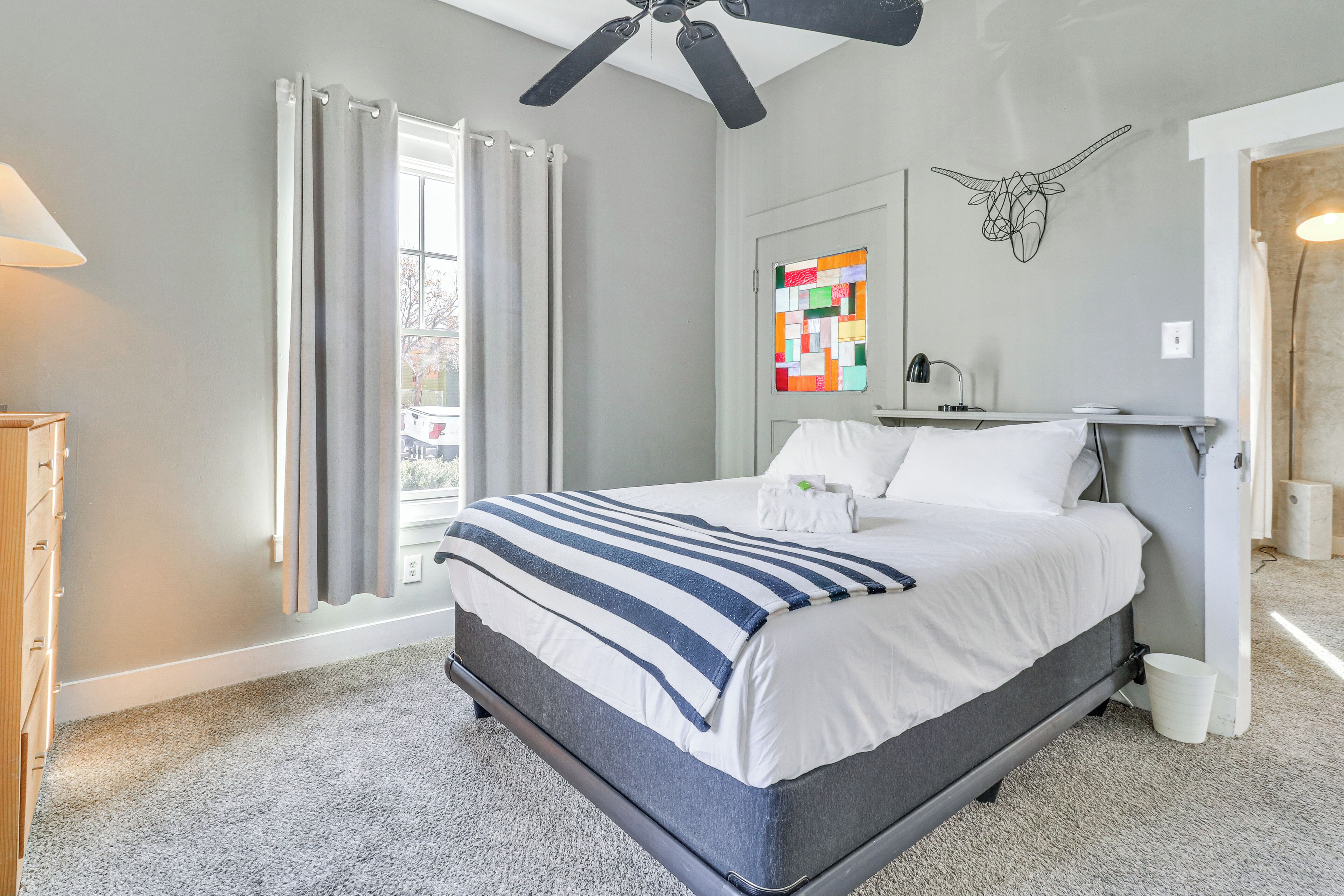 A bedroom with a bed, a ceiling fan, a window with curtains, a wall-mounted artwork, and a bedside table with a lamp.