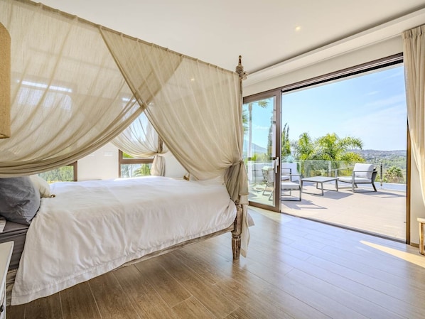 A bedroom with a four-poster bed, a balcony with outdoor furniture, and a view of the landscape.