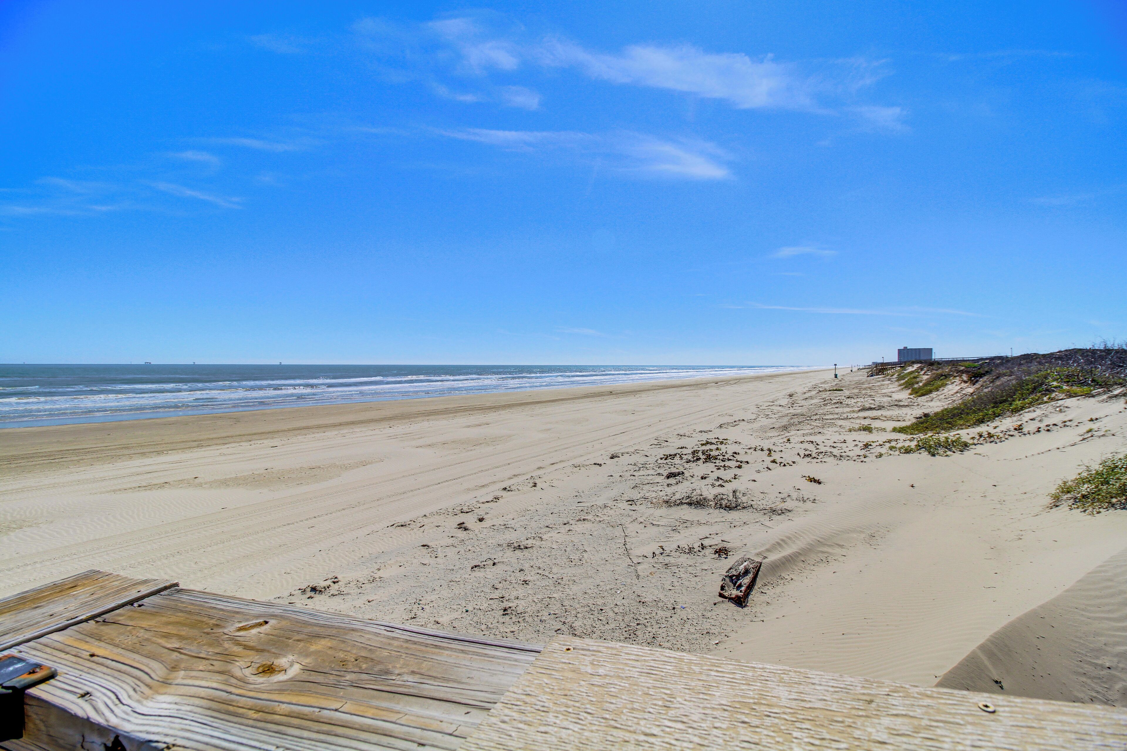 A beach with a wooden walkway leading to the shore, a sandy beach, and a distant building.