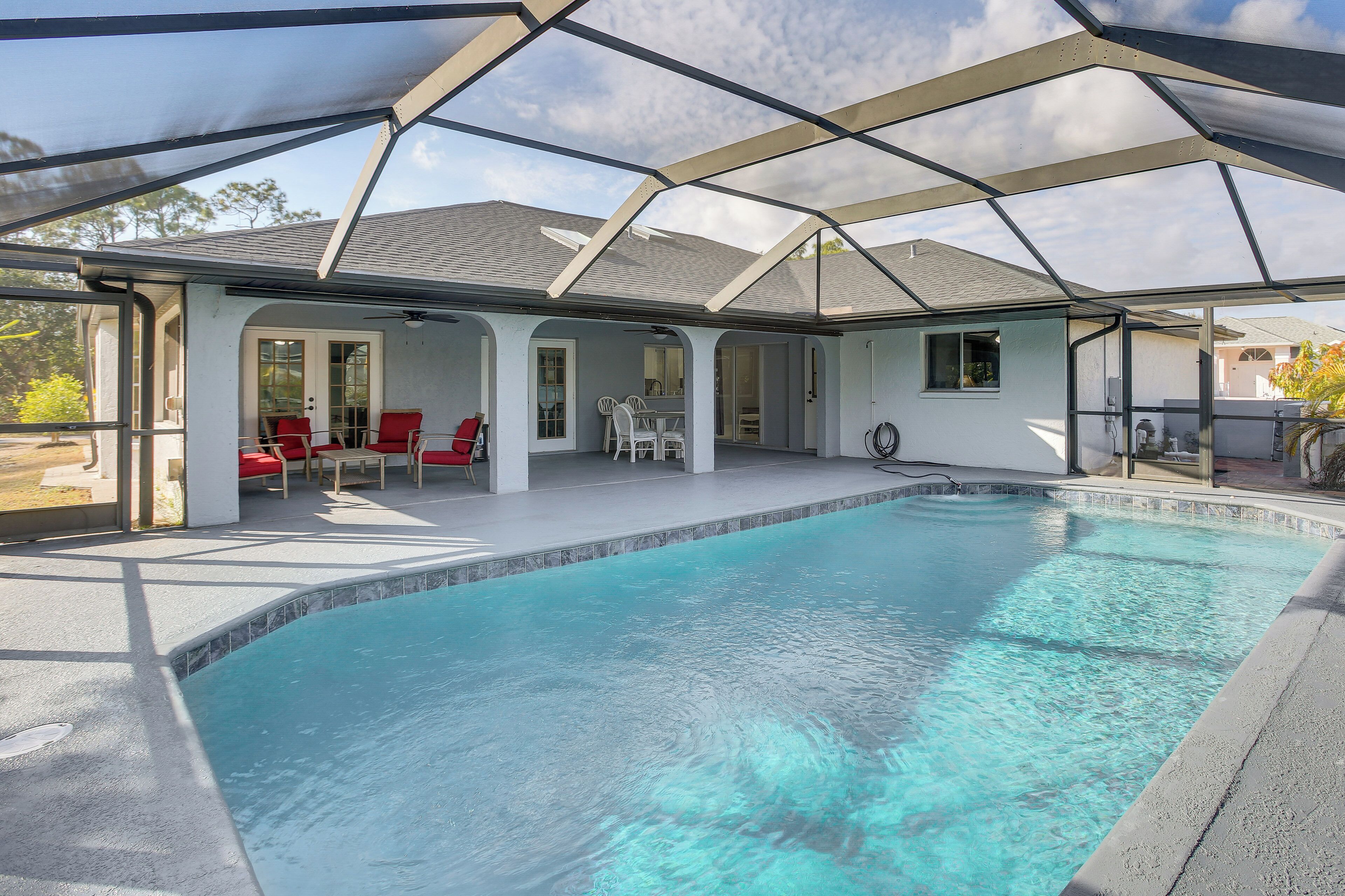 A covered swimming pool with red chairs and a table, surrounded by a house with a white exterior and a sloped roof.
