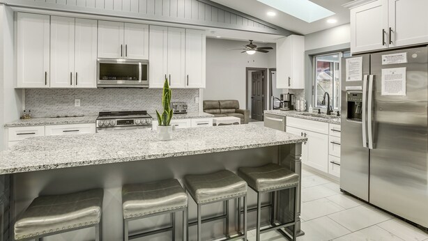 A modern kitchen with white cabinets, a stainless steel refrigerator, a large island with bar stools, and a ceiling fan.