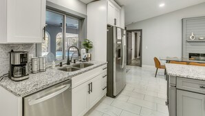 A modern kitchen with white cabinets, a stainless steel refrigerator, a coffee maker, and a window overlooking a backyard.