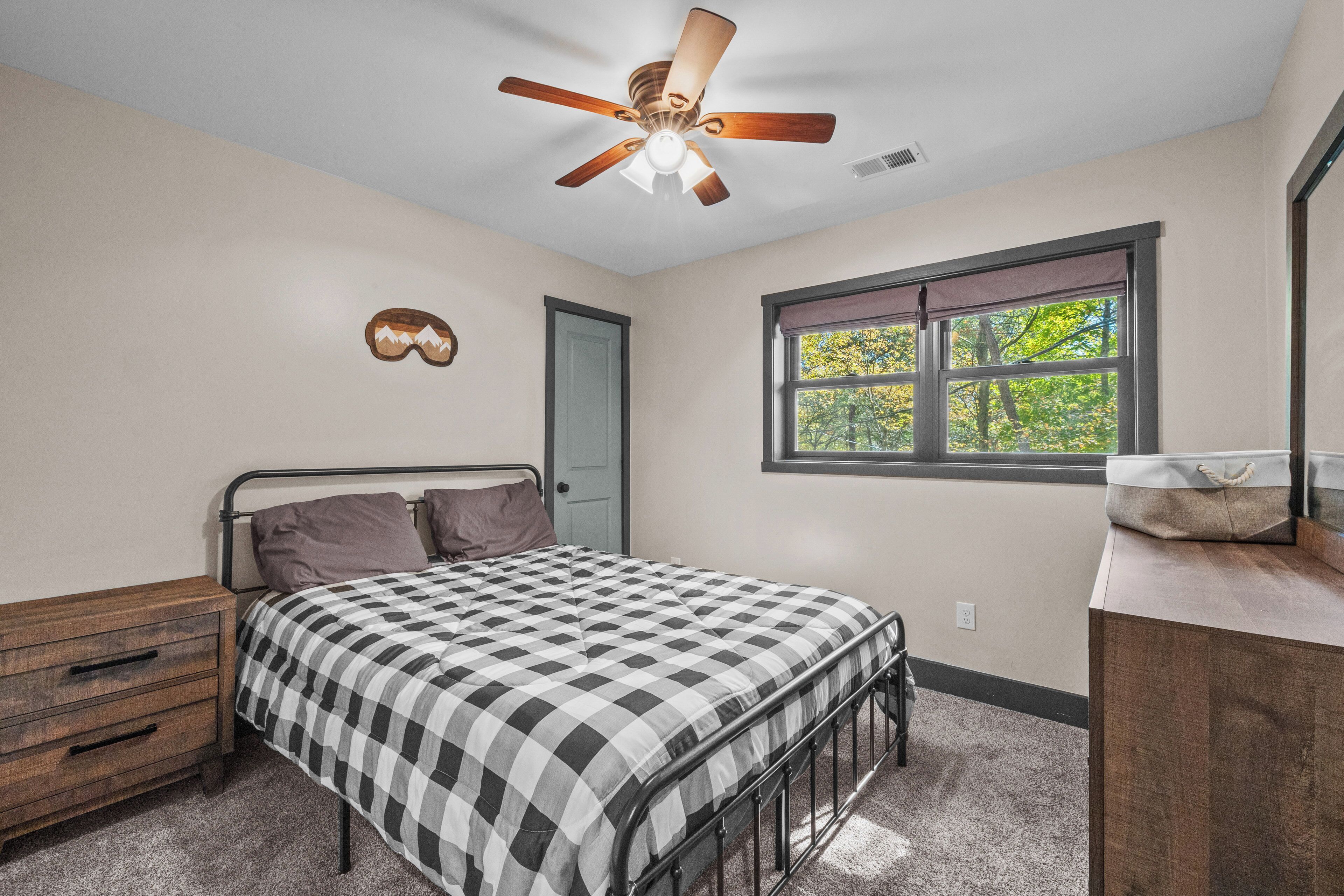 Une chambre à coucher comprenant un lit, une commode, une fenêtre donnant sur un paysage agréable, un ventilateur de plafond et une porte.