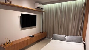 A modern hotel room with a large bed, a flat-screen TV mounted on a wooden console, a built-in air conditioner, and a large window with curtains. - Cozy NEW apartment in Ondina (Salvador)